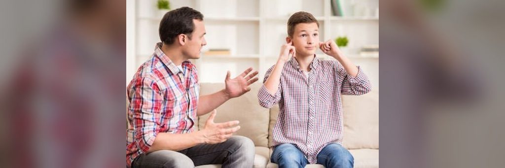 Father talks to his son on a sofa; the boy covers his ears, looking away. (in a living room)