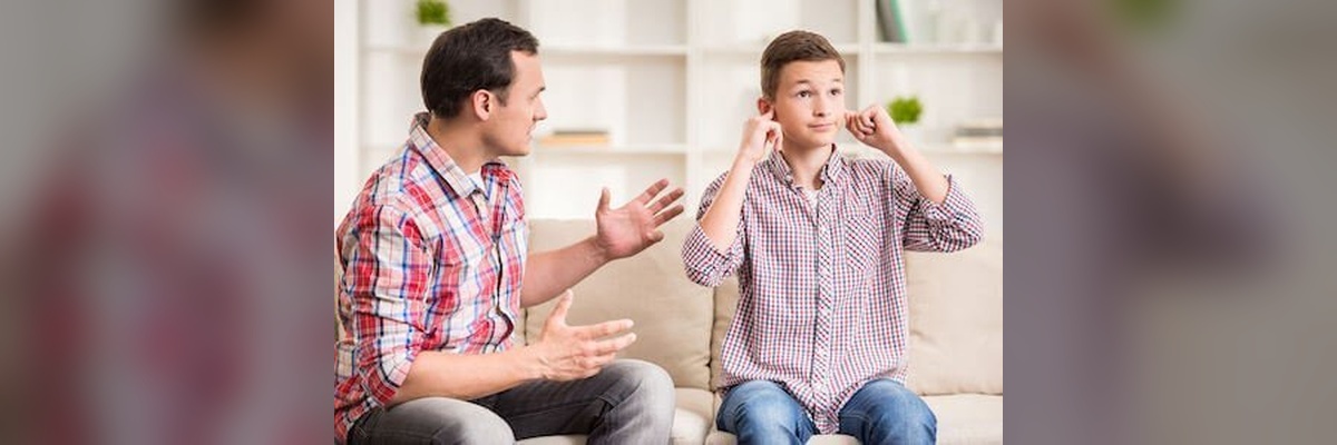 Father talks to his son on a sofa; the boy covers his ears, looking away. (in a living room)