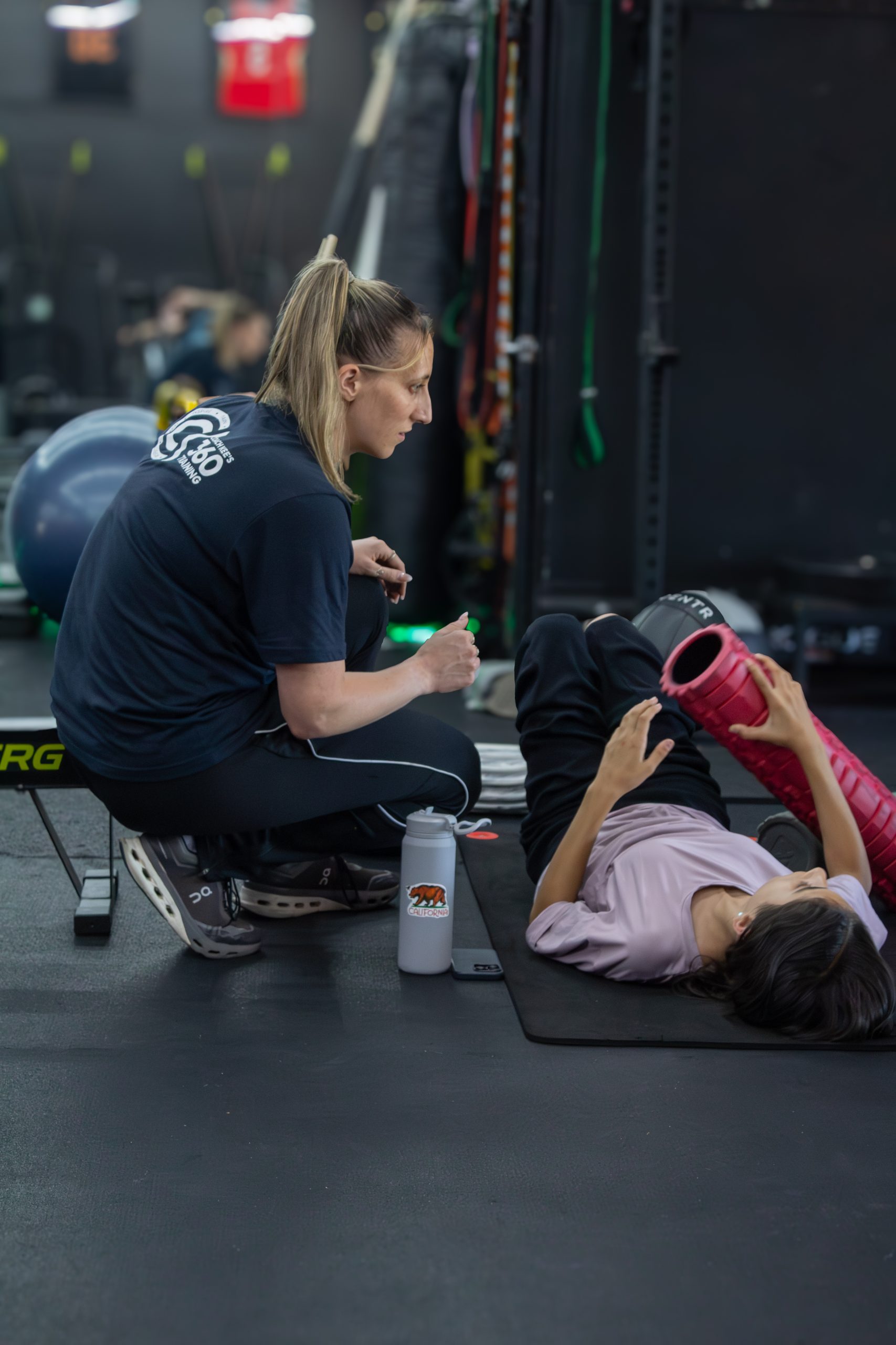Coach in a blue shirt helps a young man in a black shirt during a kneeling workout with a medicine ball nearby in a gym setting